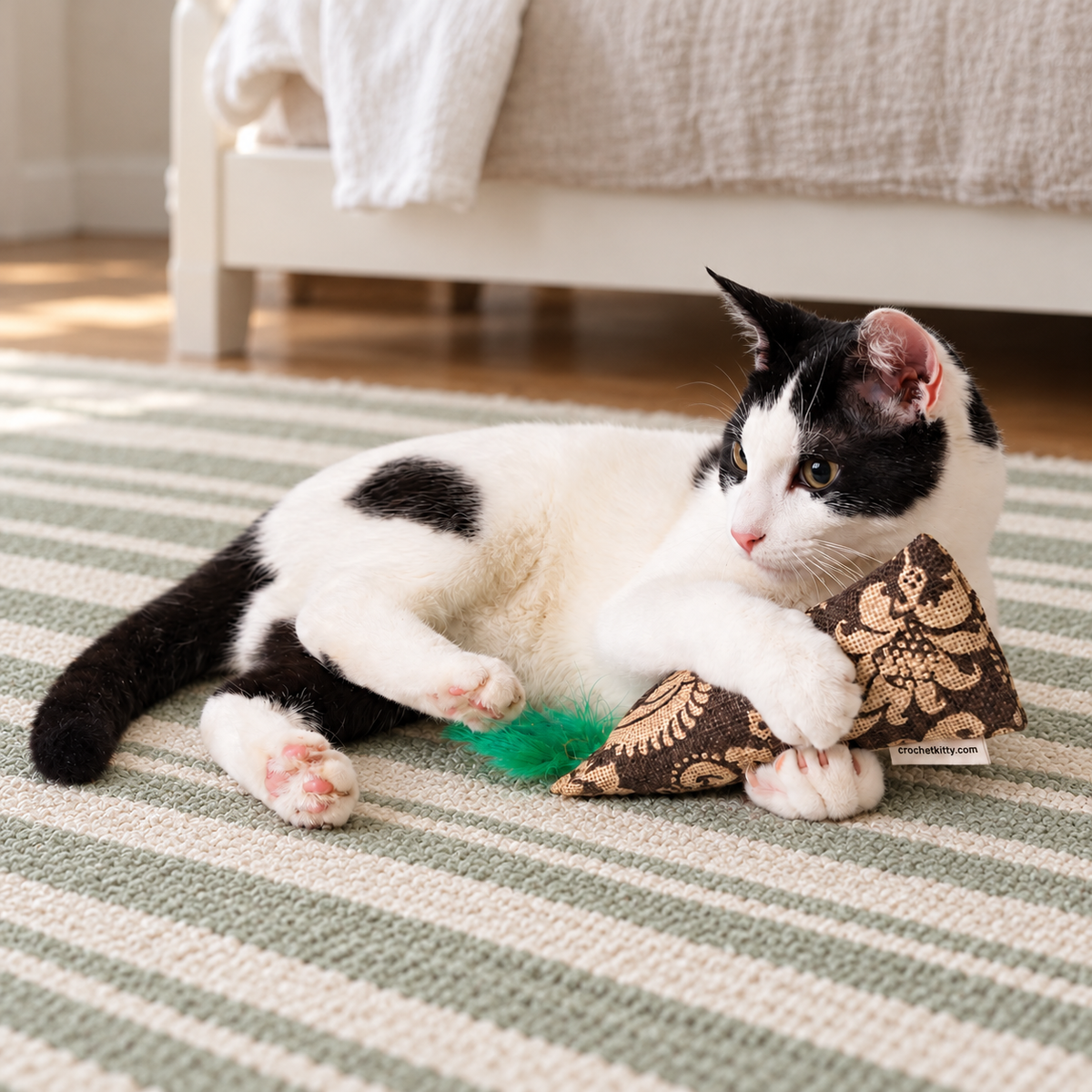 A white and black cat holding a burlap kicker cat toy with a green feather on the end. The cat is laying on a light green and beige stripped rug with a bed in the background.