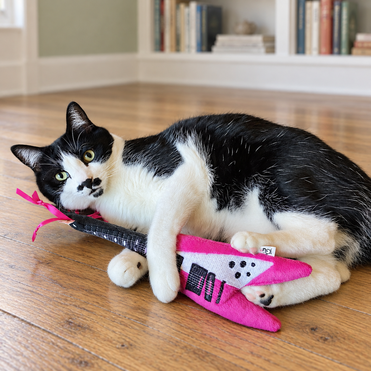 Tuxedo cat on its side hugging and kicking a pink guitar cat toy on a wood floor in a living area.