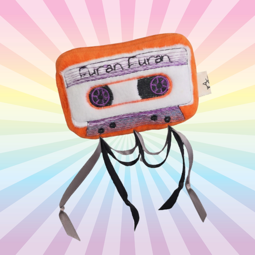Orange Furan Furan cassette catnip toy with purple embroidered details and dangling ribbons on a rainbow background.