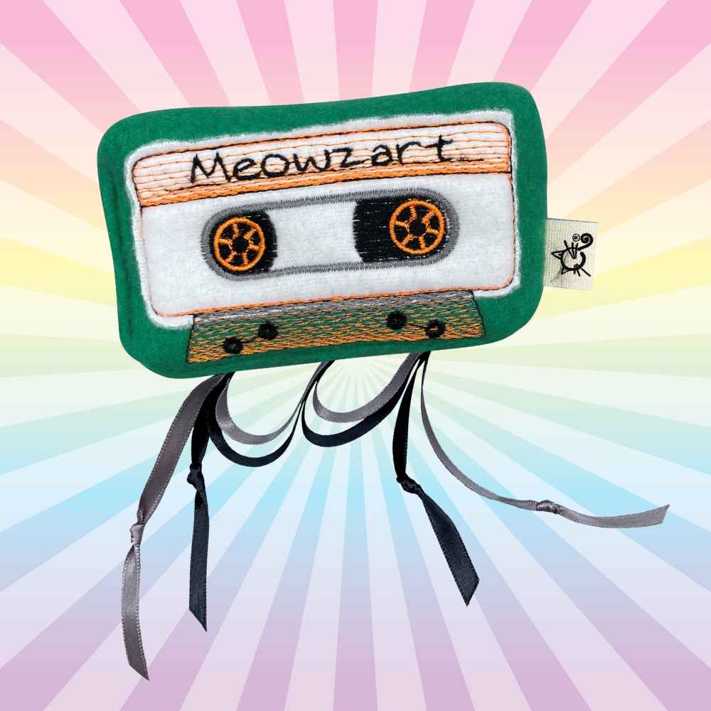 Green Meowzart cassette catnip toy with orange embroidered details and dangling black ribbons on a rainbow background.