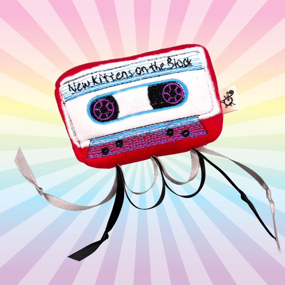 Red New Kittens on the Block cassette catnip toy with blue and purple embroidery and dangling ribbons on a rainbow background.