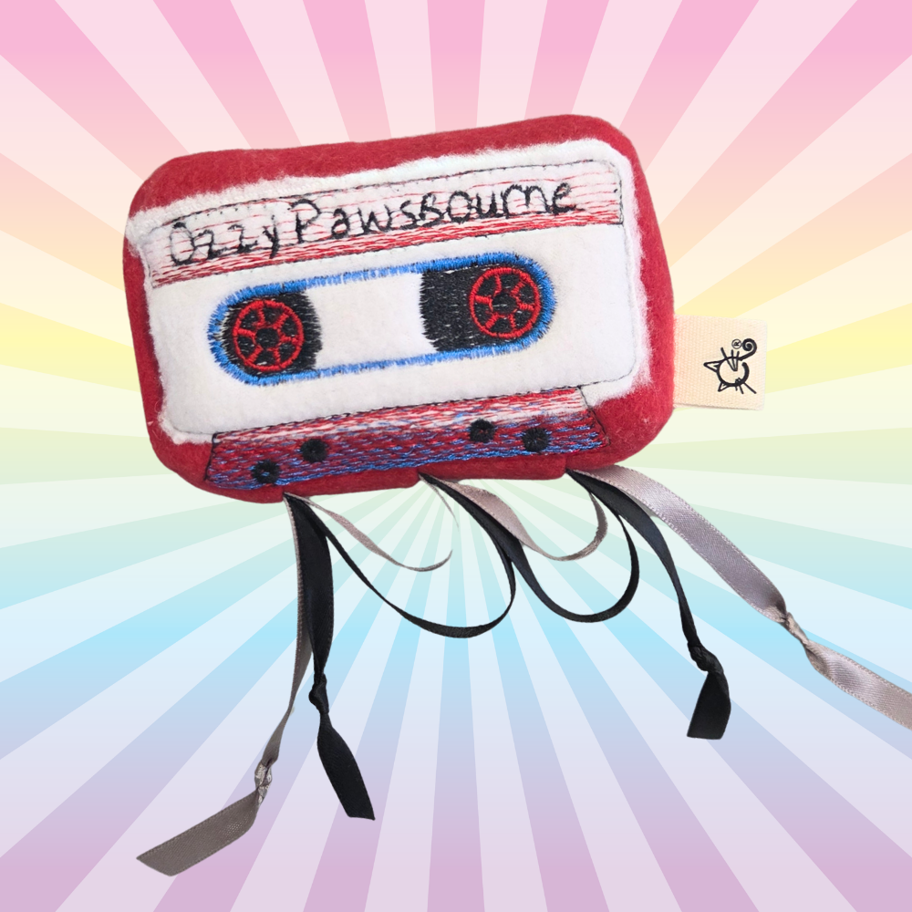 Burgundy Ozzy Pawsbourne cassette catnip toy with red embroidered reels and dangling black ribbons on a rainbow background.