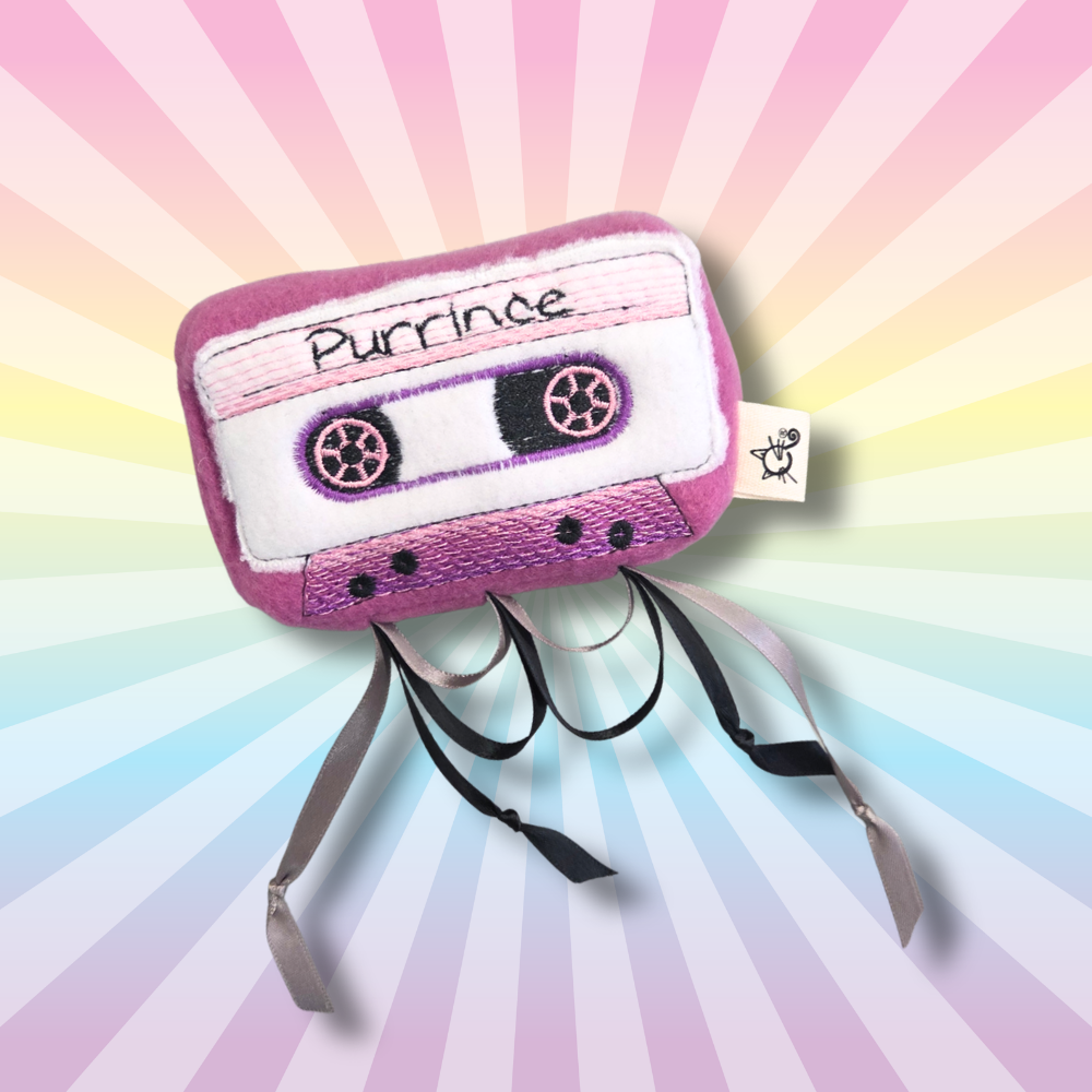 Purple Purrince cassette catnip toy with embroidered reels and dangling black ribbons on a rainbow background.
