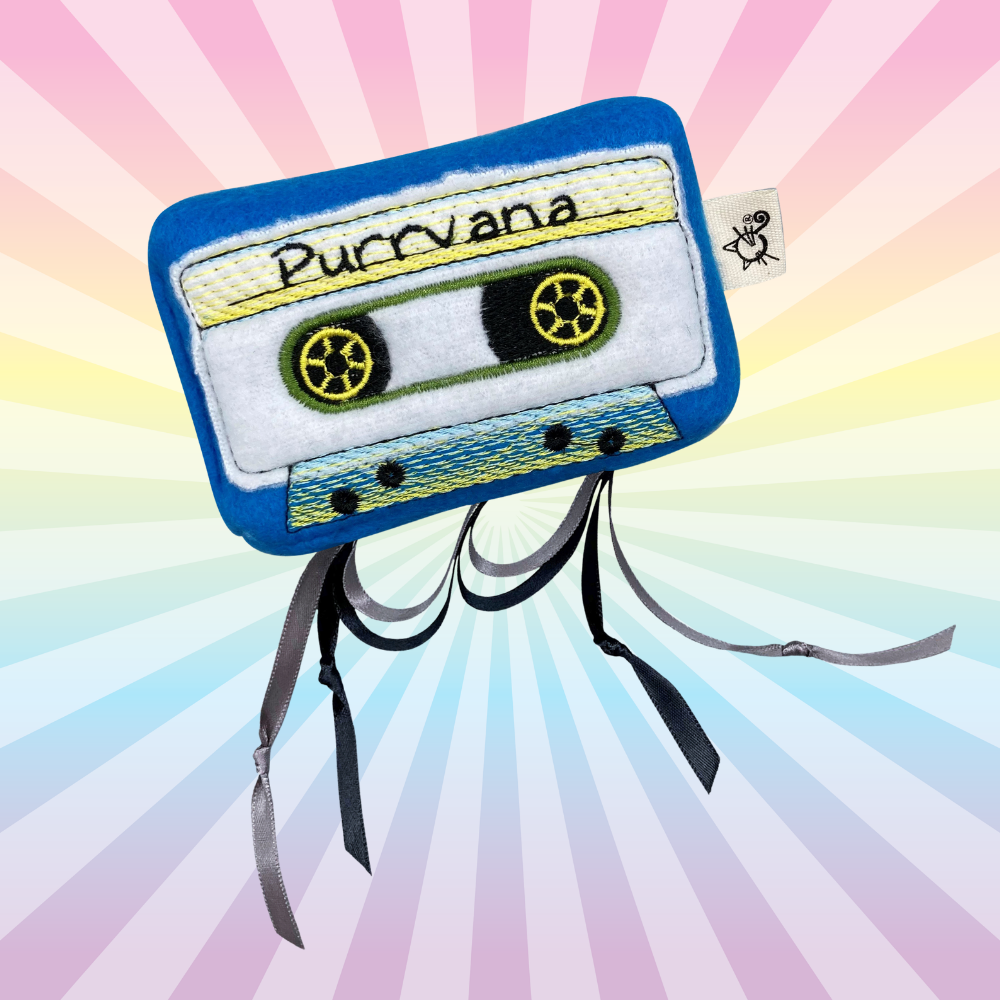 Blue Purrvana cassette catnip toy with yellow embroidered details and dangling black ribbons on a rainbow background.