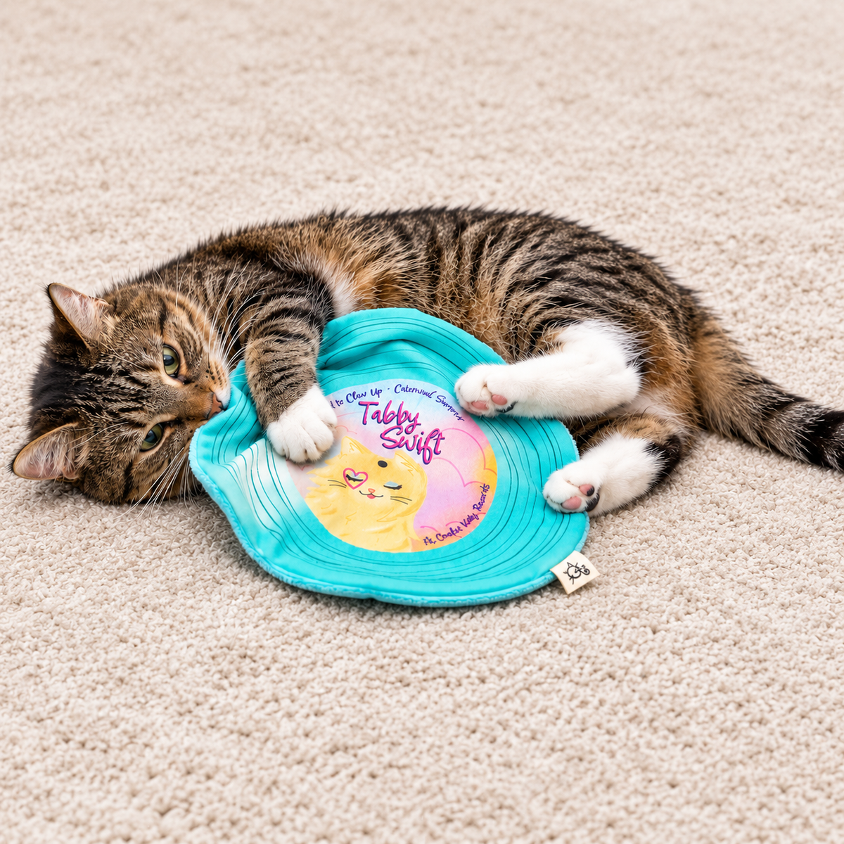 A tabby cat lies on a beige carpet, playfully biting and rolling over a bright turquoise pet toy with a yellow and pink center label that reads 'Tabby Swift'.