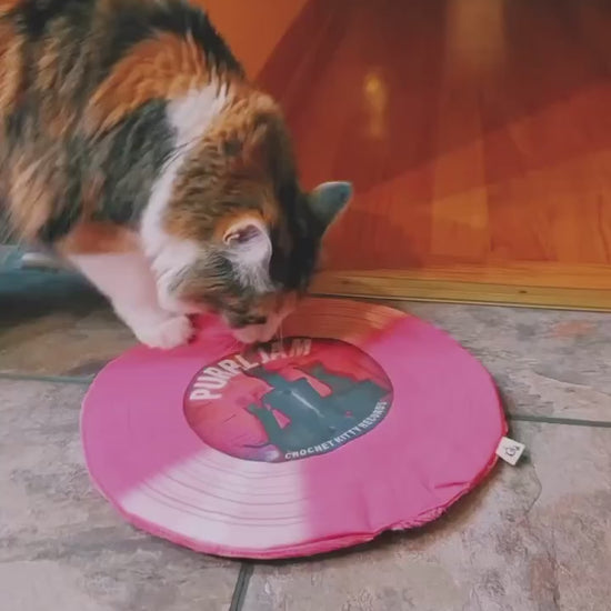 Calico cat pawing and playing with a pink Purrl Jam catnip record mat on a tiled floor.