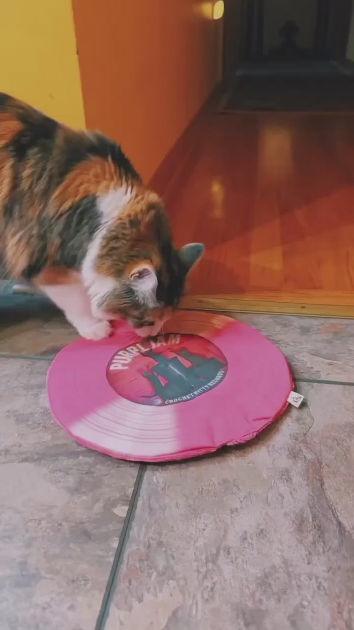 Calico cat pawing and playing with a pink Purrl Jam catnip record mat on a tiled floor.